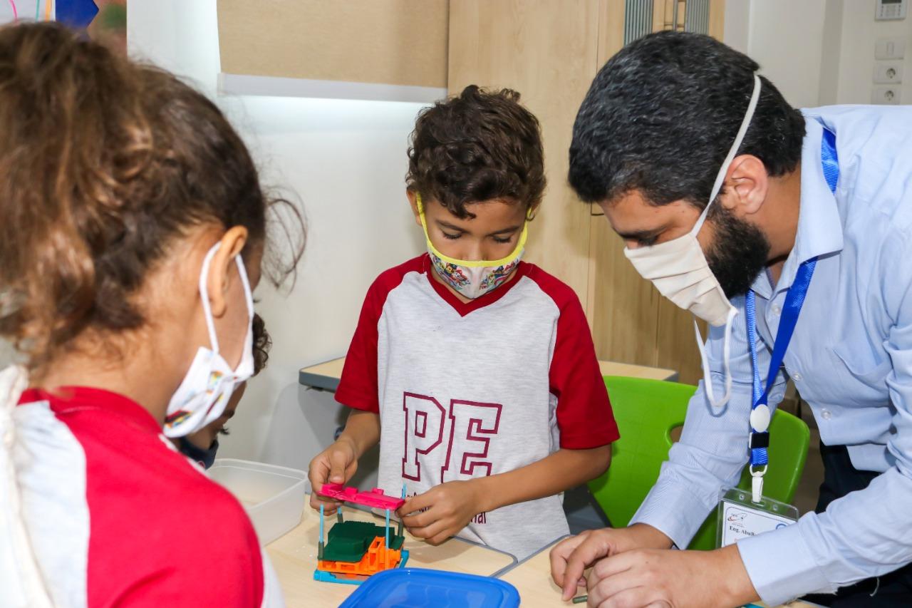 A teacher at IVY STEM International School engages with students in a hands-on learning activity involving colorful building blocks. The classroom setting promotes problem-solving, construction, and design skills.