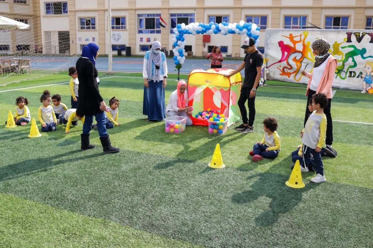 A group of young children wearing yellow tops, accompanied by adults, participating in an outdoor event organized by IVY STEM International School. The event showcases a playful atmosphere with a colorful play tent, bright plastic balls, and yellow cones for organized activities. The banners in the background depict dancers and children in motion, indicating a theme related to children's play, sports, or dance.