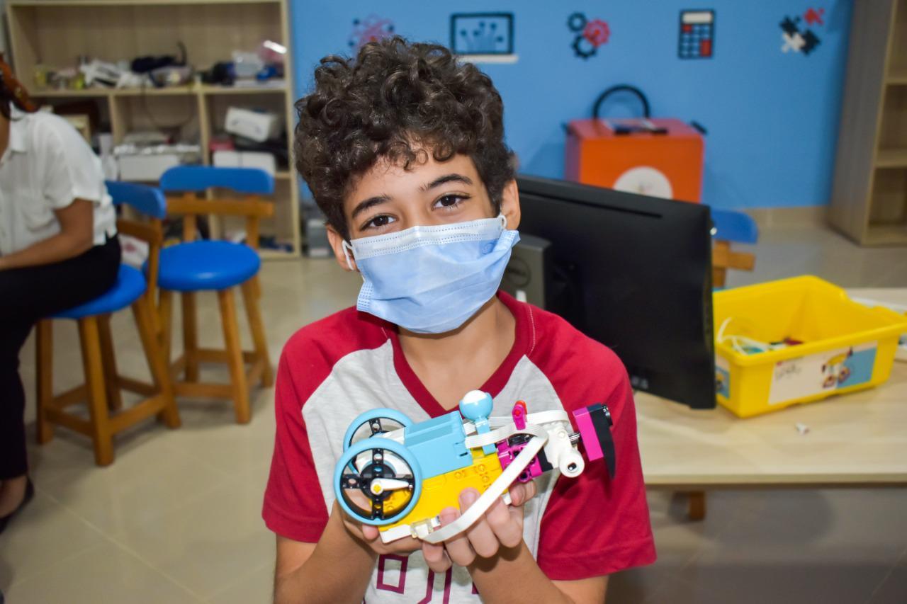 A young person with curly hair holding up a colorful STEM learning project resembling a vehicle made of LEGO blocks at IVY STEM International School.